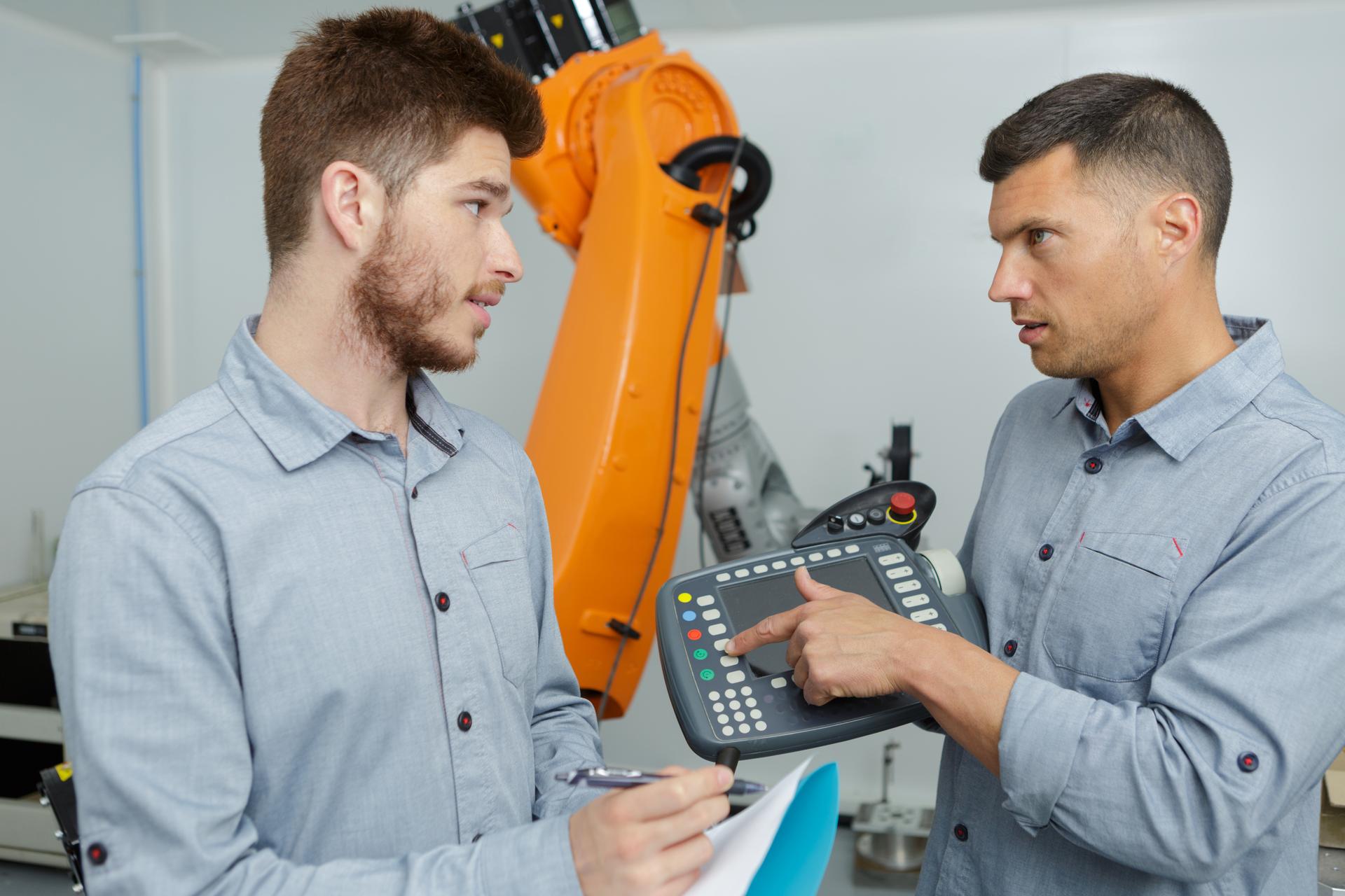 to personer af hankøn kigger på hinanden, i baggrunden ses en orange robotarm 